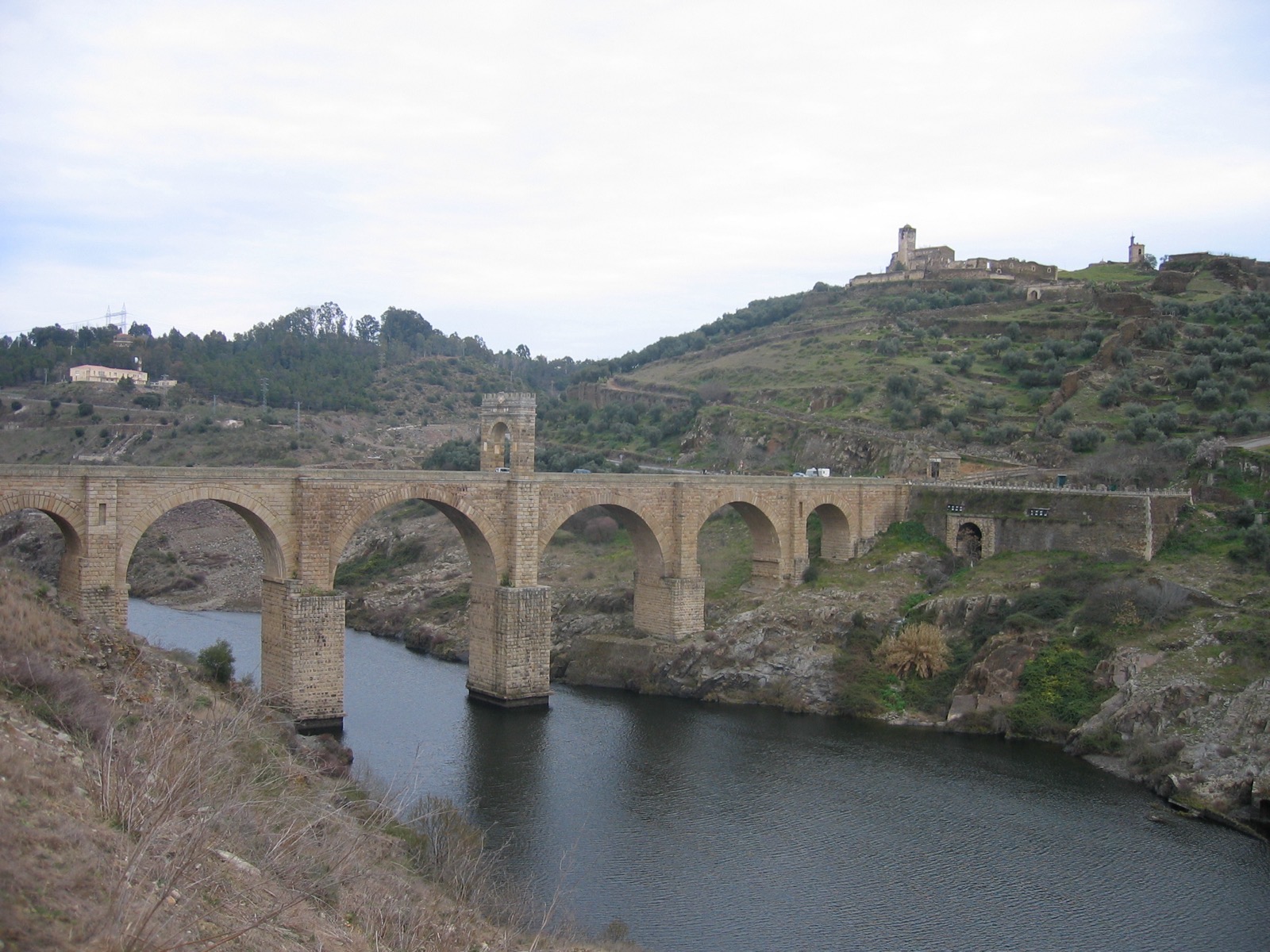 The Alcántara Bridge, a Roman stone arch bridge over the Tagus River in Spain, showing its full six-arch span with the central triumphal arch tower and a hilltop temple in the background
