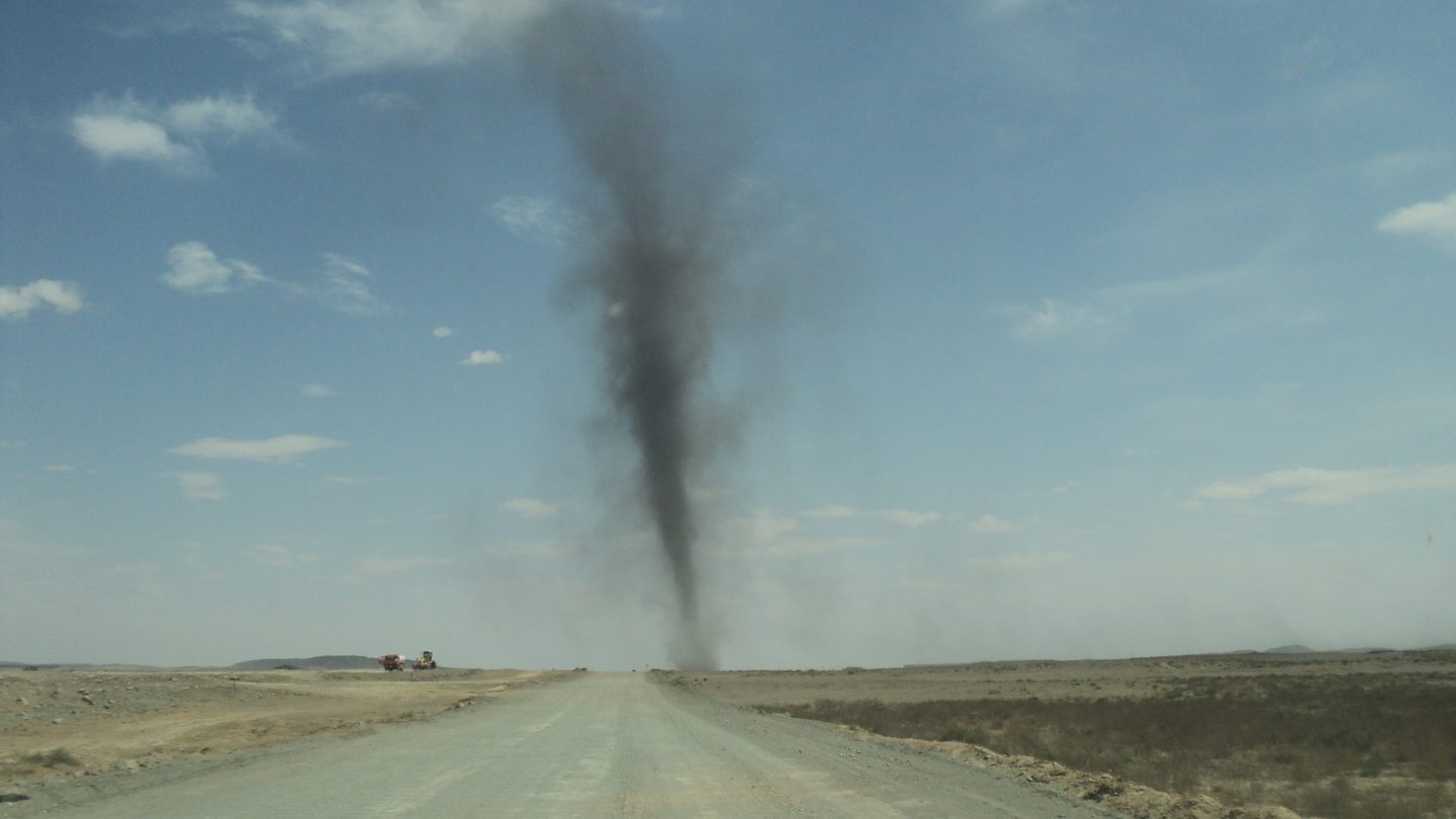 A tall dark column of dust and coal particles whirling above a dirt road in Mongolia, with small vehicles visible in the distance