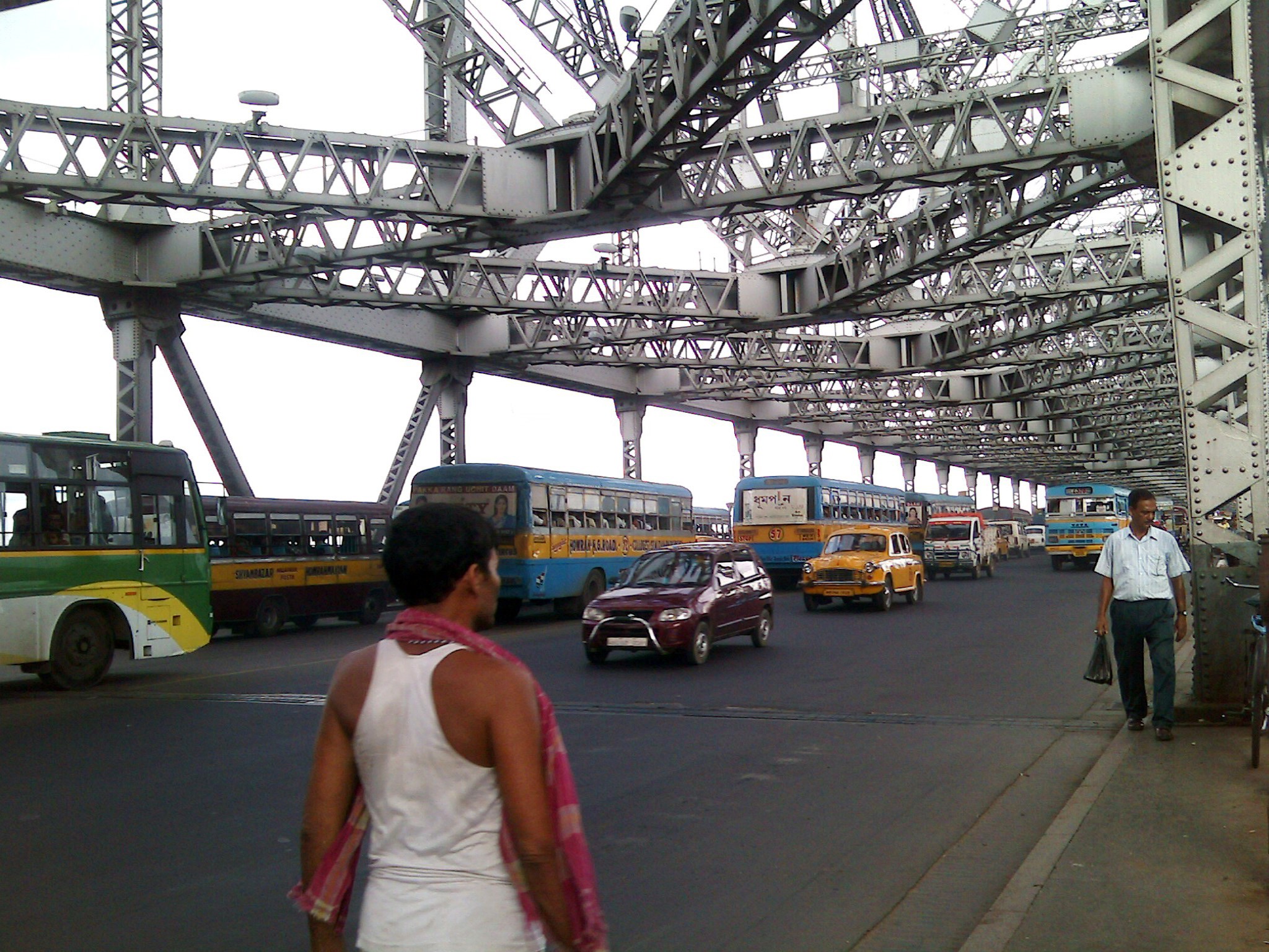 Traffic crossing Kolkata's Howrah Bridge over the Hooghly River, showing the riveted steel cantilever structure and the dense stream of vehicles and pedestrians it carries daily