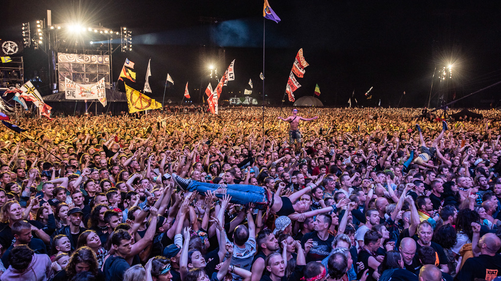 A dense festival crowd at night under stage lights, arms raised and flags waving as one person is passed overhead above the crowd