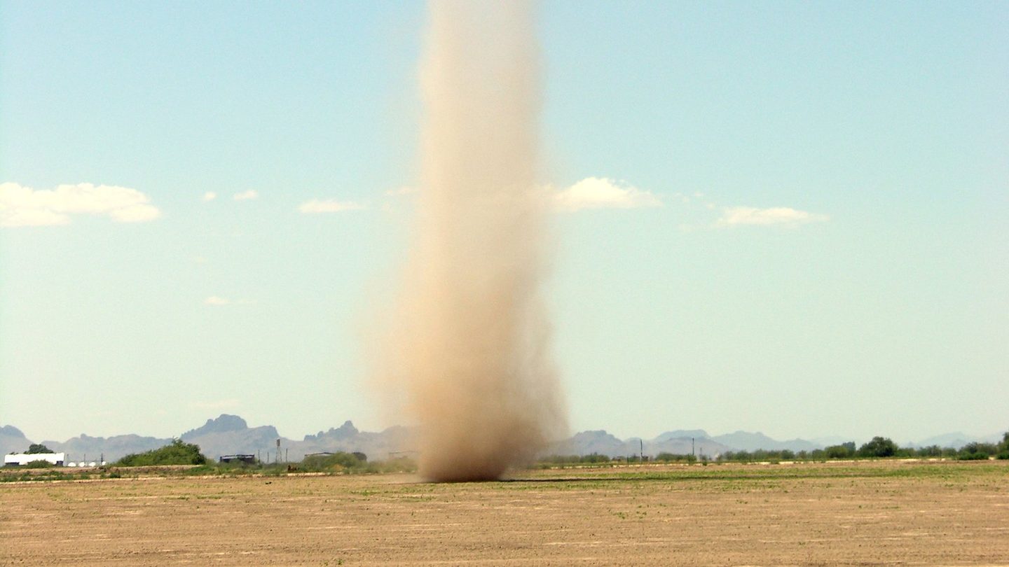 A tall column of brown dust rising from a flat dry field in Arizona under a clear blue sky with only a few small clouds, a dust devil with no storm cloud above it
