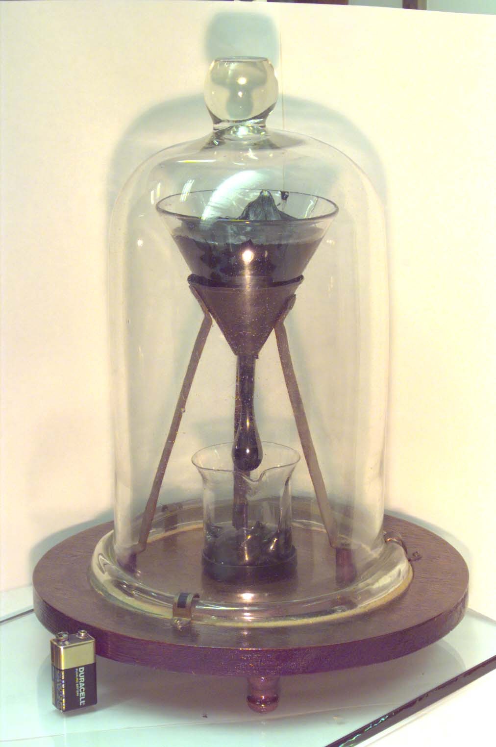 The University of Queensland's pitch drop experiment: a dark lump of pitch inside a sealed glass funnel, photographed with a 9-volt battery for scale