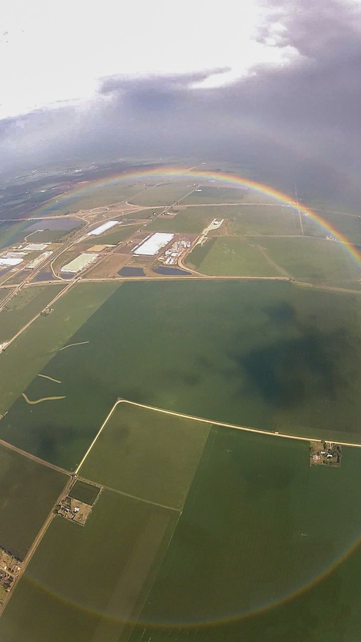 Aerial view of green farmland from a skydiver's height with a rainbow forming a nearly complete circle around the viewpoint, the upper arc bright and the lower part faint